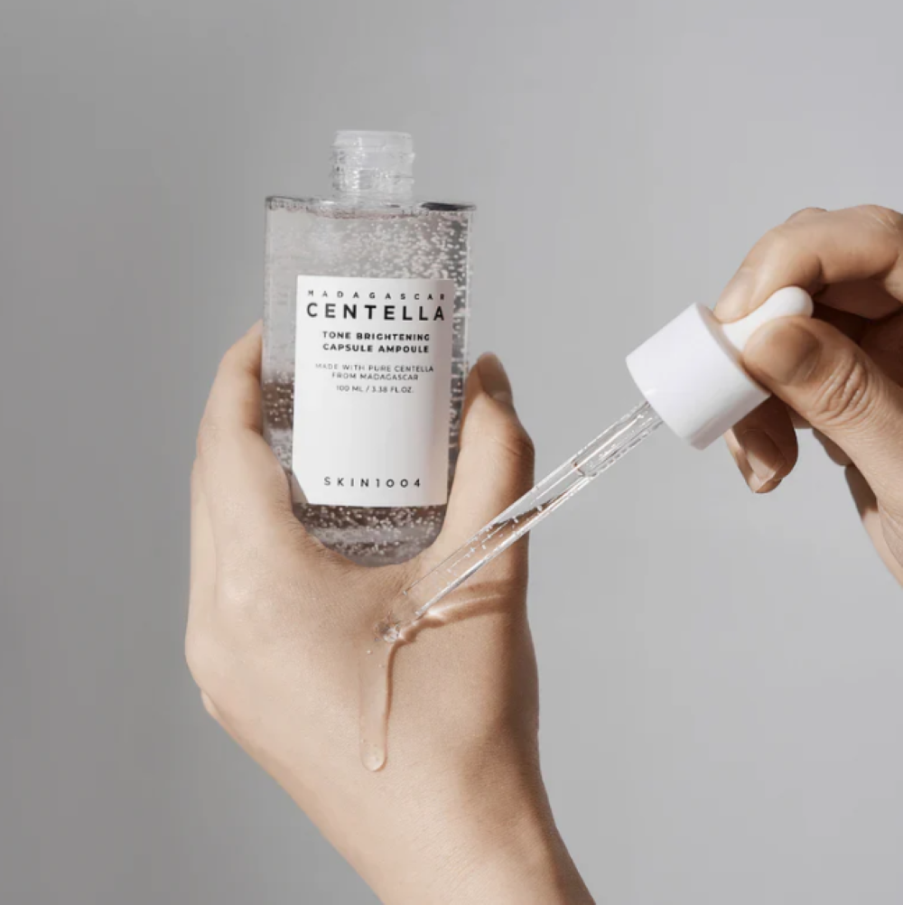 Hand holding a skincare bottle with dropper against a gray background, Skin1004 Madagascar Centella Tone Brightening Capsule Ampoule 100ml