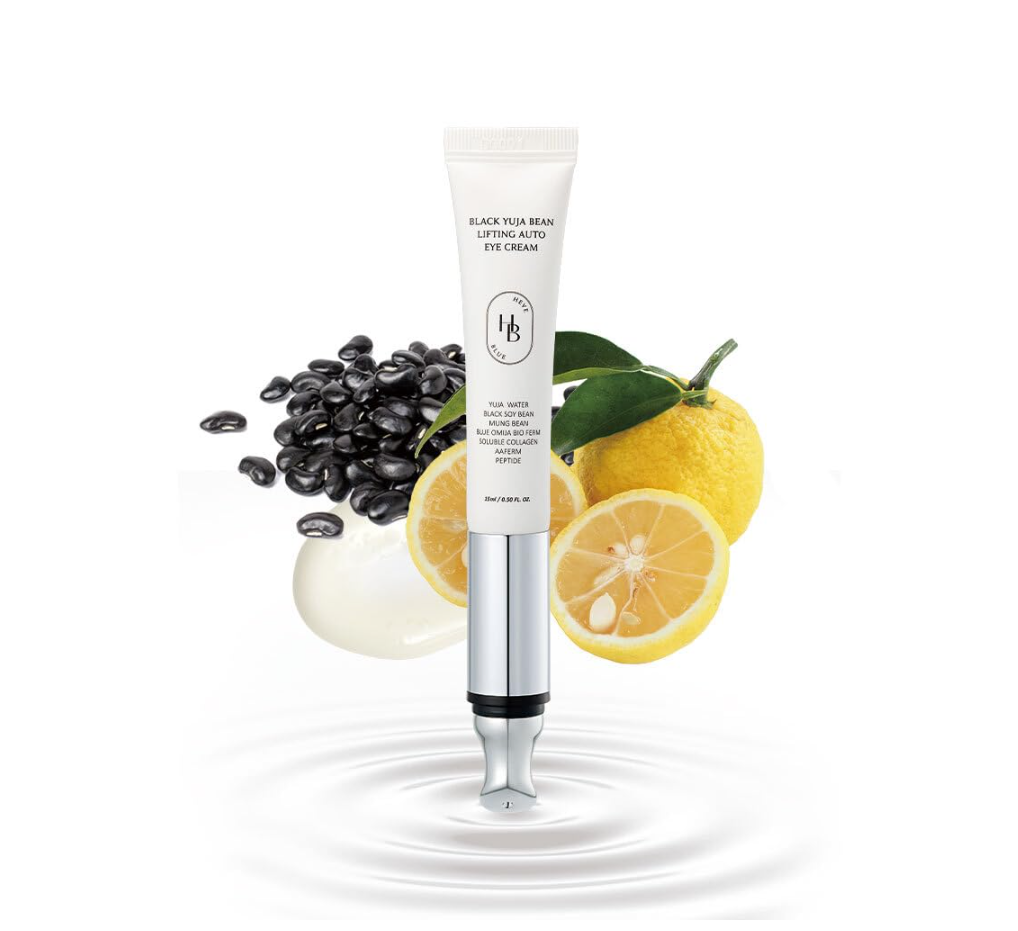 A white and silver tube of black seed eye cream is centered on a white background, surrounded by decorative props including whole and sliced lemons with green leaves, scattered black seeds, and a dollop of cream with a ripple effect at the bottom. Heveblue Black Yuja Bean Lifting Auto Eye Cream 15ml