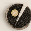 A white cosmetic tube labeled 'KOKUA YU SOUL GEL CREAM' rests diagonally on a bed of black beans inside a speckled ceramic bowl, with a sliced lime half as a prop on a light beige background. Heveblue Black Yuja Bean Lifting Auto Eye Cream 15ml