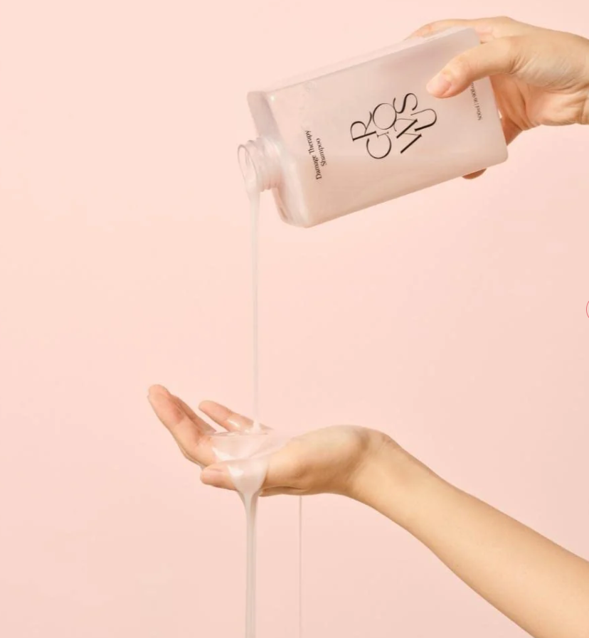 Person pouring a white liquid from a bottle labeled 'BCS' onto their hand against a pink background, Growus Damage Therapy Shampoo EX 500ml